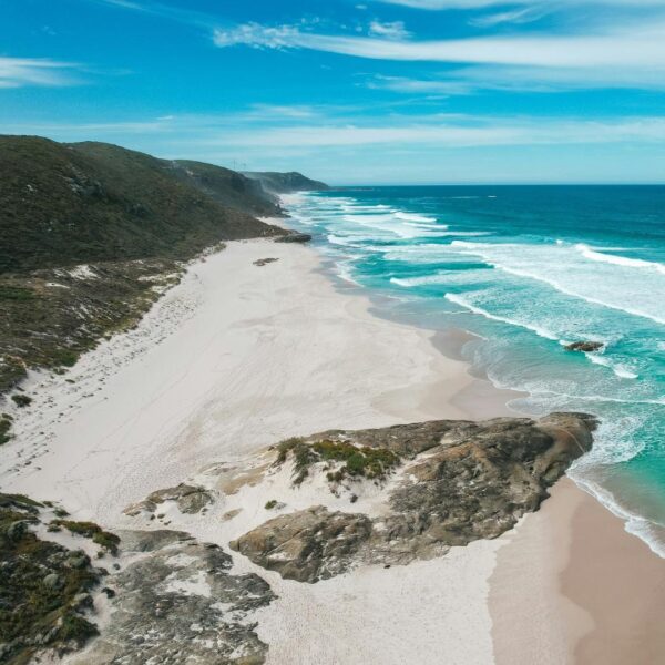 Long stretch of sand interrupted by large rocks with green ocean waves with white spray lapping at the sand